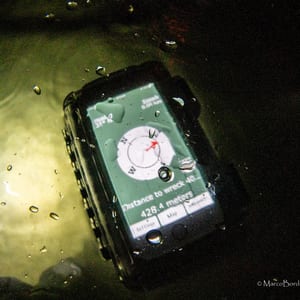 A handheld unit in a waterproof case underwater, its green screen showing a compass rose and the reading Distance to wreck 428.4 meters.