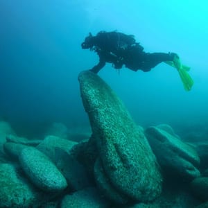 A scuba diver in a dark wetsuit with bright yellow fins hovers just above the tip of a tall pointed boulder rising from a bed of rounded rocks, in clear green-blue water.