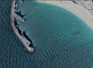 Aerial view of a curved stone breakwater reaching out from a sandy beach, with dark blue deep water on one side and pale turquoise shallow water inside the bay, where wave lines fan around behind the breakwater arm.
